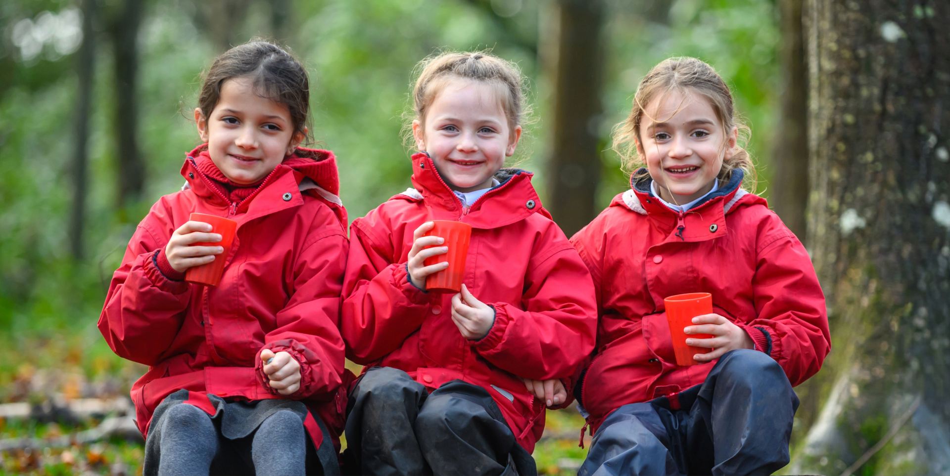 three students in red jackets in the woodland holding up red cups