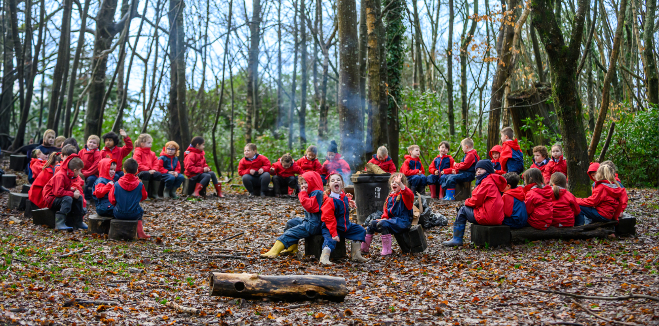 Students in red jackets sitting in a circle in the woods for a Forest School lesson.