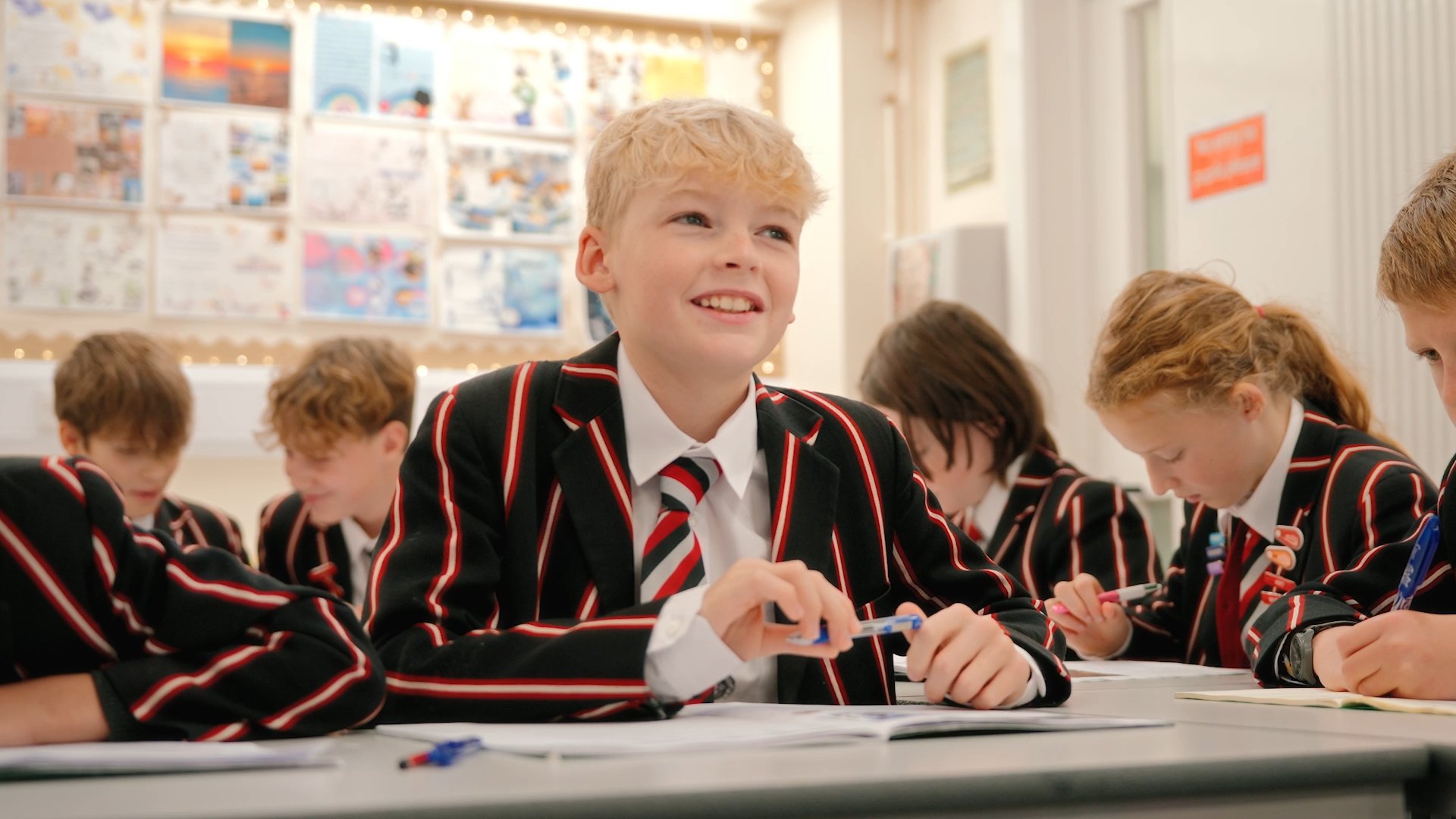 A smiling student in a striped school blazer sitting in a bright classroom.