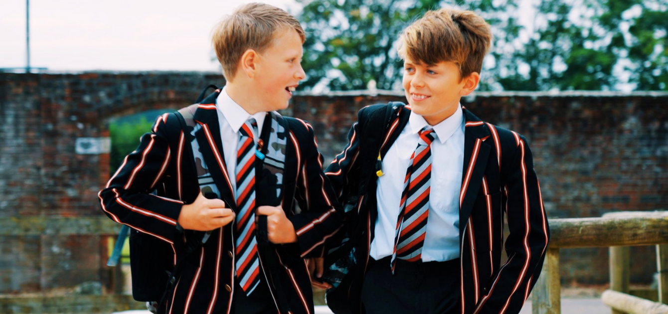 Two boys in striped school blazers and ties walking and talking outdoors.